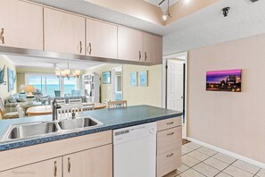 A modern kitchen with a large island, stainless steel sink, and white cabinetry. There is a dining area with a view of the ocean and a living room with a fireplace. - Sea Gate 303 - GULF FRONT Condo! (Indian Shores)