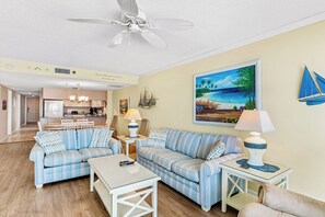A living room with striped sofas, a ceiling fan, a painting of a beach, and a sailboat decoration. - Sea Gate 303 - GULF FRONT Condo! (Indian Shores)