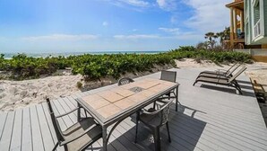 Une terrasse en bois avec une table et des chaises, donnant sur une plage bordée de palmiers et sur laquelle on aperçoit un bâtiment au loin.