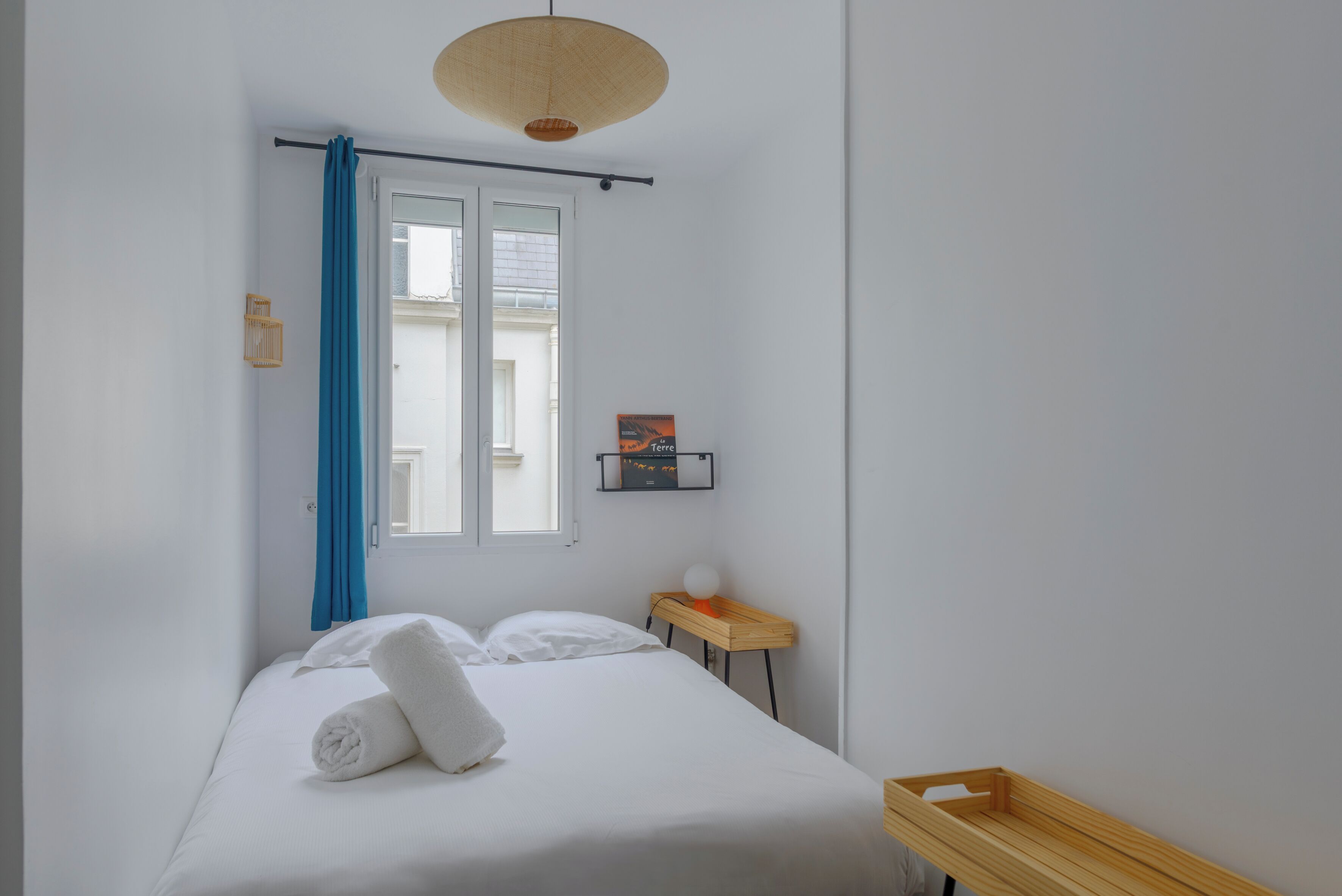 A neatly made bed with white linens and a rolled towel, a small wooden side table with a spherical object, a wall-mounted shelf with a book, and a window with blue curtains.