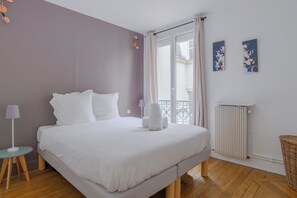 Quarto com cama, mesa de cabeceira, abajur, radiador e vista para a varanda.