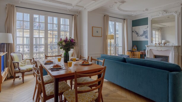 A dining area with a wooden table, chairs, and a blue sofa. There is a window with curtains and a view of buildings outside. - Paris sacré coeur prestige residence (Paris)