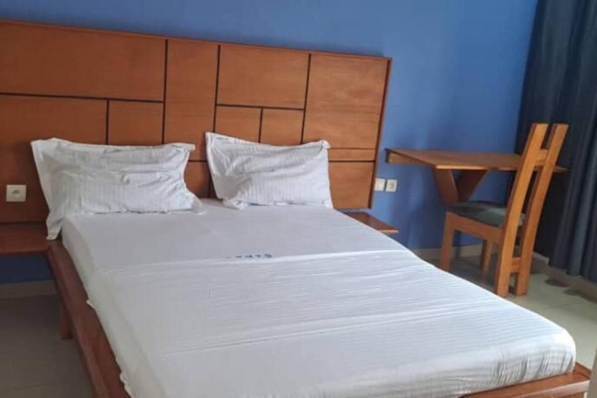 Hotellihuone, jossa on puinen sängynpääty, valkoisilla lakanoiden peitetty sänky, pieni puinen pöytä ja tuoli.