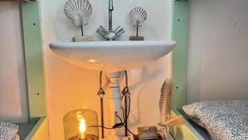 A compact room with a bed, a small shelf with decorative items, a wall-mounted radiator, and a bedside table with a lamp.