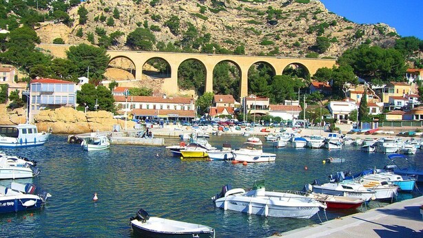 Uma marina com barcos atracados, uma ponte de pedra e edifícios ao fundo.