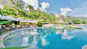 A resort pool with lounge chairs and umbrellas, surrounded by palm trees and buildings. - Magnificent Ocean view suite with private balcony jacuzzi (Puerto Vallarta)