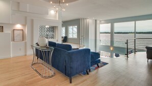 A modern living room with a large window overlooking a body of water, a blue sofa, a glass coffee table, and a unique wall art piece.
