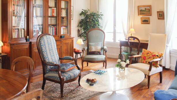 A well-furnished living room with a wooden bookshelf, a round coffee table, and various seating options including a blue armchair and a floral armchair. - Timeless apartment in a Haussmannian building (Paris)