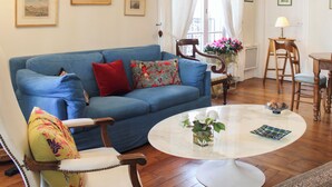 A living room with a blue sofa, a round coffee table, and a floral-patterned chair. - Timeless apartment in a Haussmannian building (Paris)