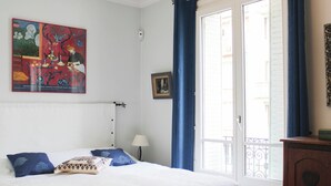 A bedroom with a bed, a nightstand, a lamp, a window with curtains, and a painting on the wall. - Timeless apartment in a Haussmannian building (Paris)
