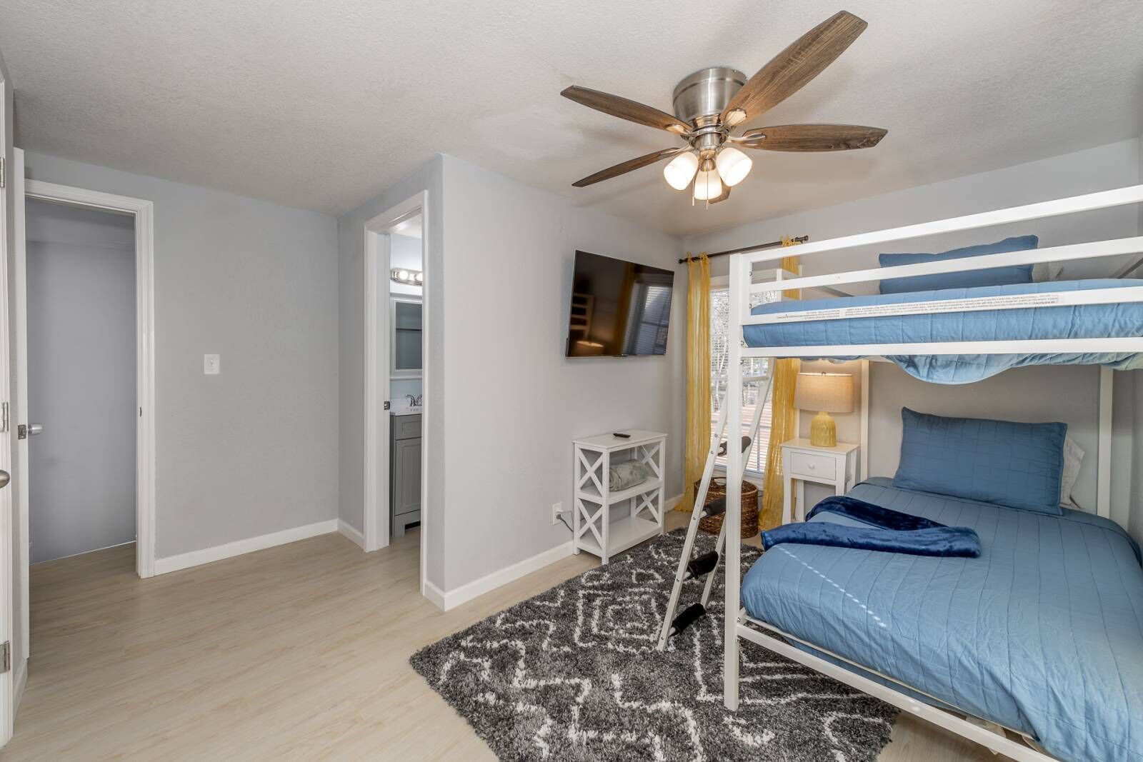 A bedroom with a bunk bed, a ceiling fan, a desk, and a small window with curtains.