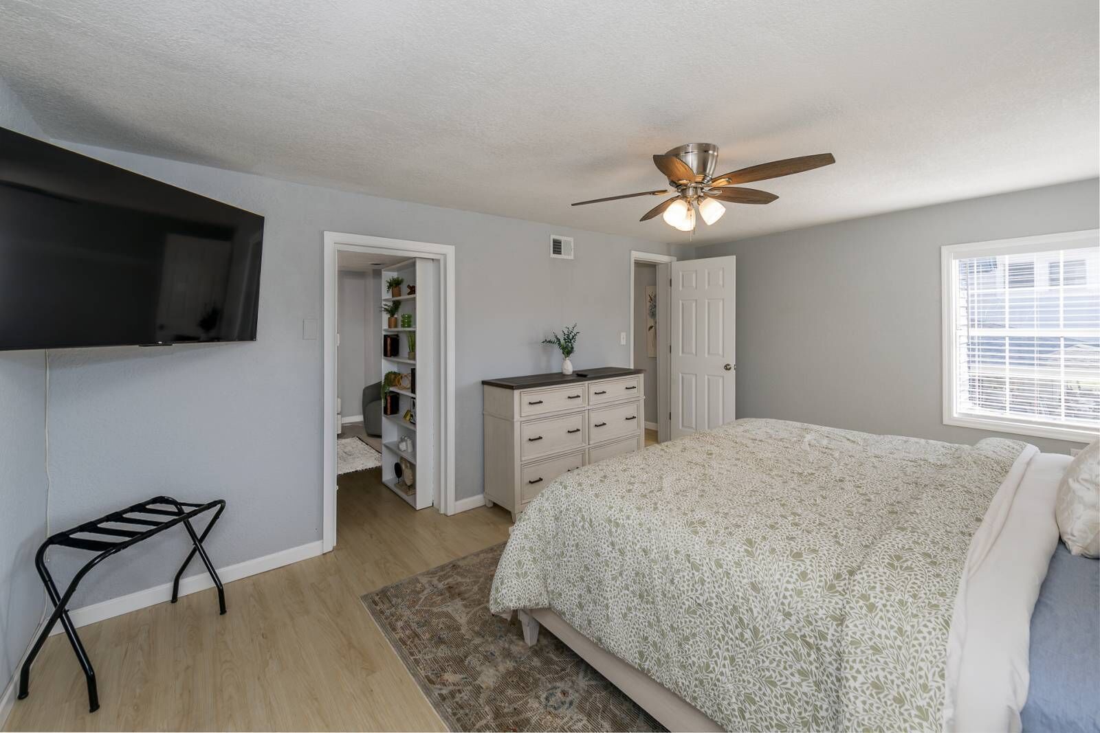 Une chambre à coucher comprenant un lit, une commode, un téléviseur fixé au mur, un ventilateur de plafond et une fenêtre avec des stores.