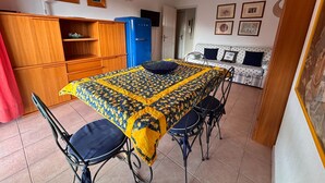 Ein Essbereich mit einem Tisch, der mit einer gemusterten Tischdecke bedeckt ist, umgeben von Metallstühlen. Im Hintergrund ist ein blauer Kühlschrank zu sehen, und eine mit gemusterten Kissen ausgelegte Couch lehnt an der Wand.