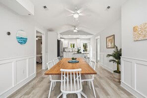 A spacious dining area with a wooden table and white chairs, a ceiling fan, and a view into the kitchen. - The Salty Fin: Dog-Friendly, Walk to Everything! (Oak Island)