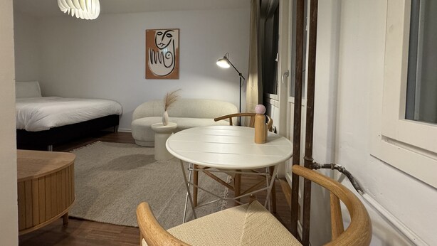 Ein gemütliches Schlafzimmer mit einem Bett, einem kleinen weißen Tisch mit Lampe, einem Sessel und einer dekorativen Vase.