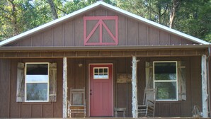 Una cabaña rústica con puerta roja, dos ventanas en la fachada y un porche de madera.