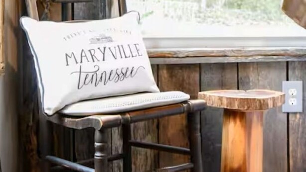 Una silla de madera rústica con un cojín que dice "MARYVILLE tennessee" junto a una ventana con vistas a árboles.