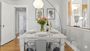 Un coin repas avec une table blanche dressée pour quatre personnes, un vase de fleurs et un luminaire suspendu moderne.