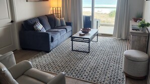 A living room with a sofa, armchair, coffee table, and a view of the beach through large windows. - Regency Towers #402W Beach Front Condo, Balcony, Pool, Pickleball , Sleeps 8 (gulfbreeze)