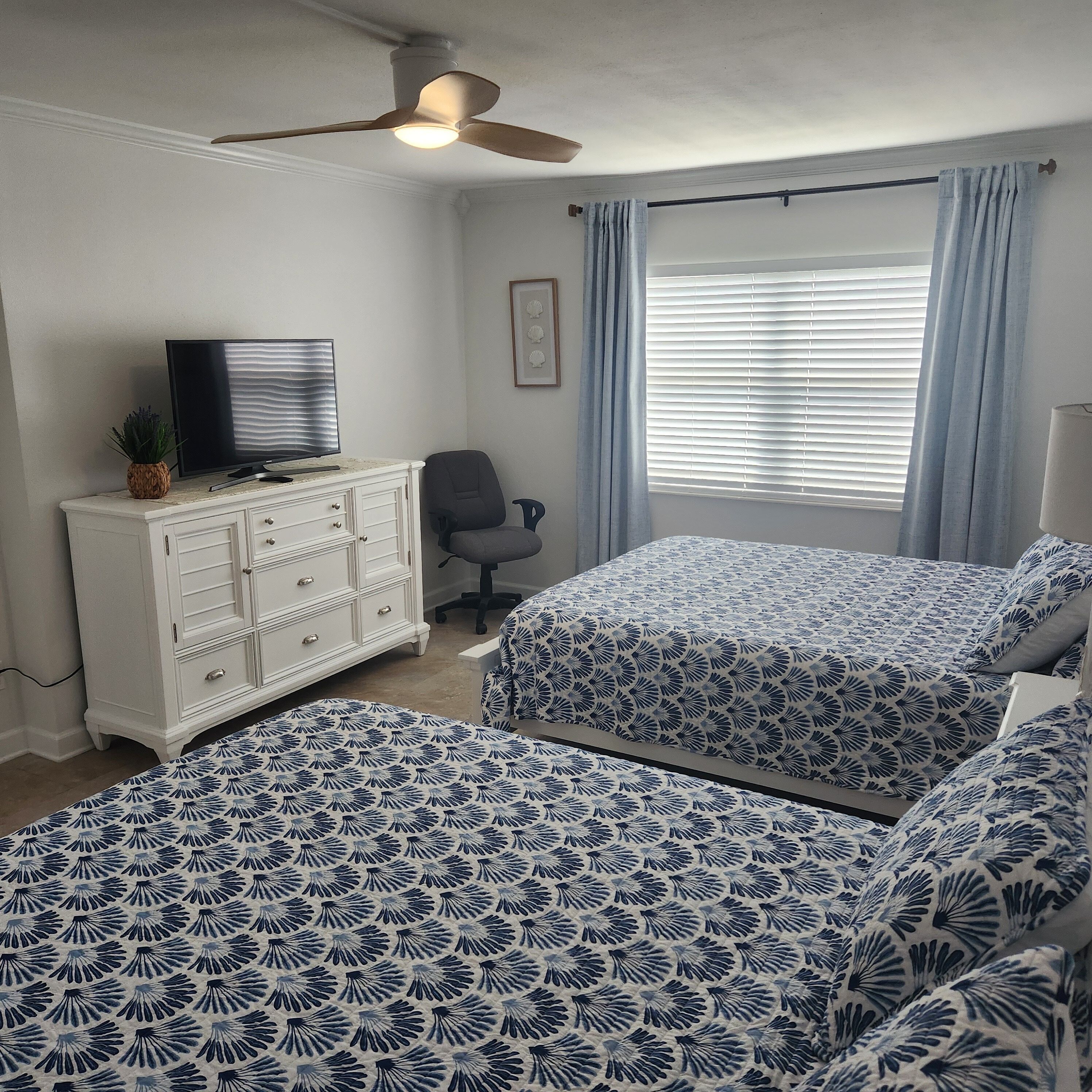 Une chambre avec deux lits, une commode, un téléviseur, un ventilateur de plafond et une fenêtre avec des rideaux.