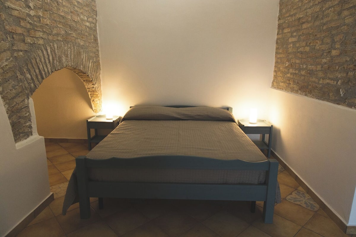A narrow room with a bed centered between two stone walls, flanked by nightstands with lamps.