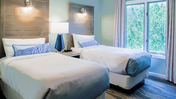 Hotellihuone, jossa on kaksi sänkyä, yöpöytä lampun kanssa, ikkuna verhoilla ja näkymä vehreään maisemaan.