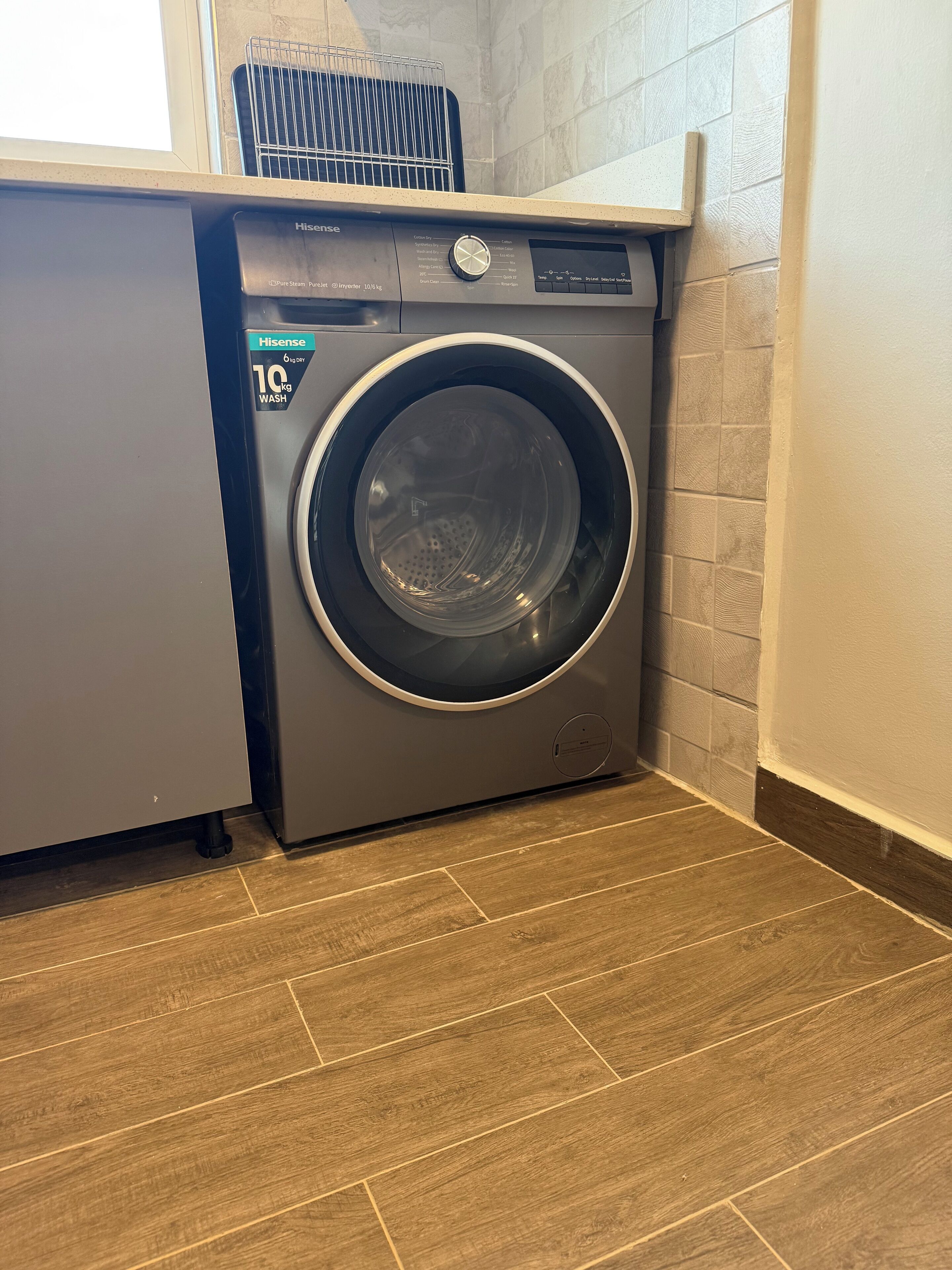A front-loading washing machine with a capacity of 10 kg wash, located in a laundry room with wooden flooring and a tiled wall.