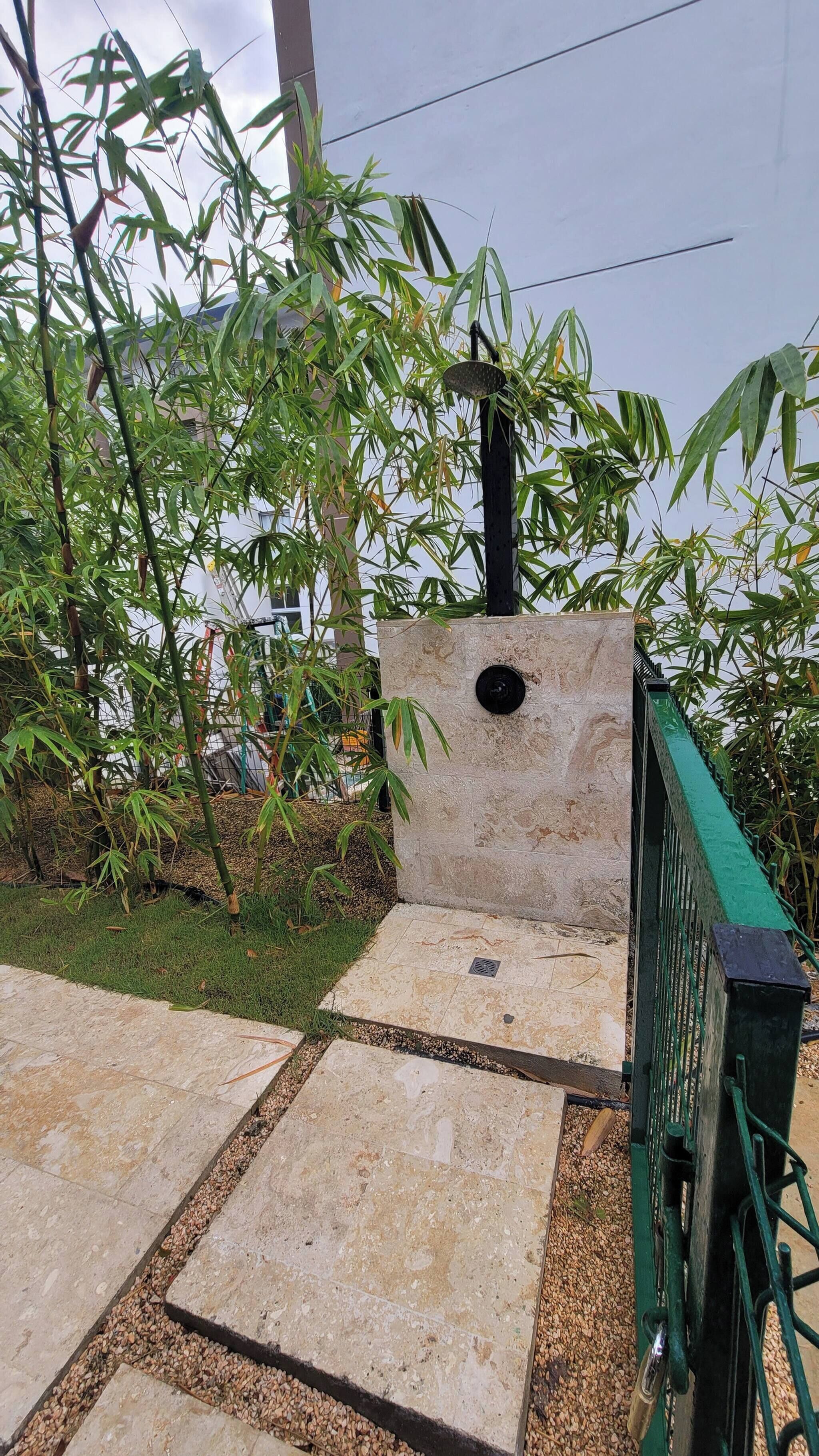 Uma trilha pavimentada conduz a uma estrutura de pedra com uma abertura circular, cercada por plantas de bambu e uma cerca de metal verde.