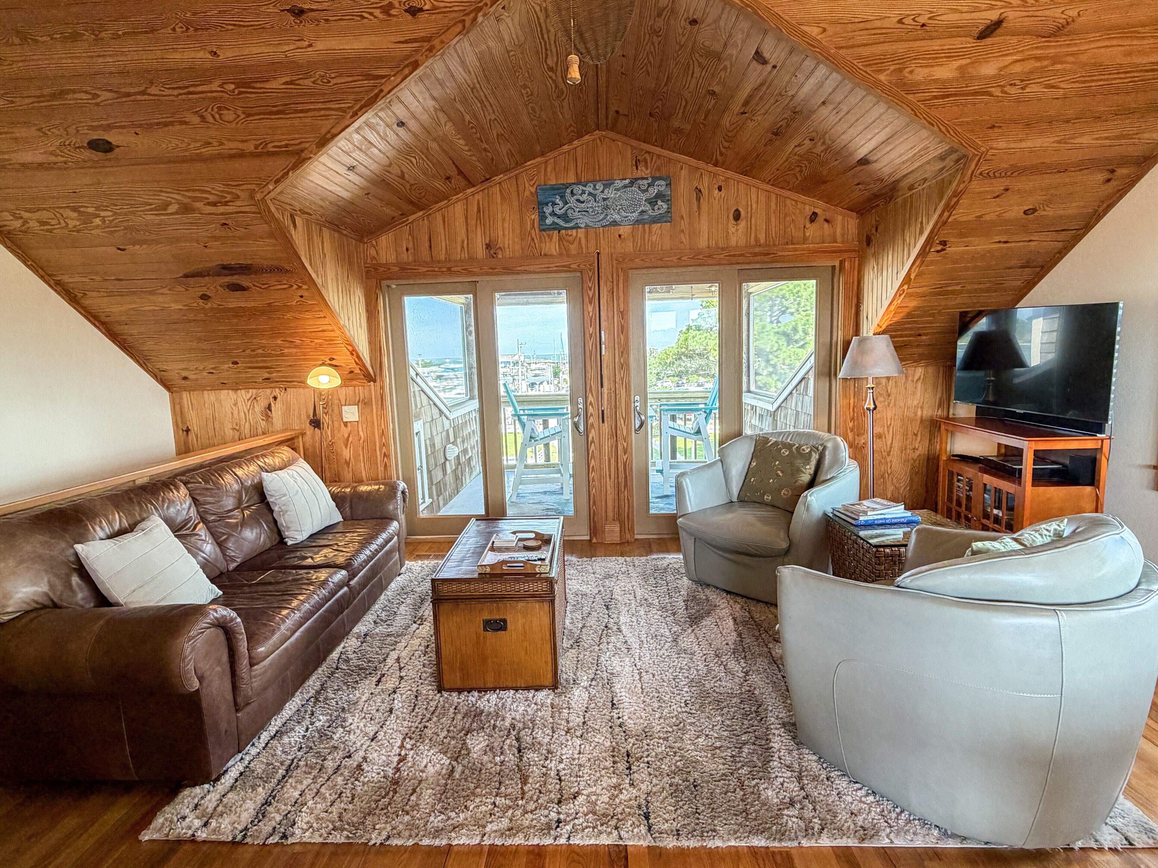 A cozy living room with a brown leather couch, a white armchair, and a wooden coffee table. A television is mounted on the wall, and there is a large window with a view of the outdoors.