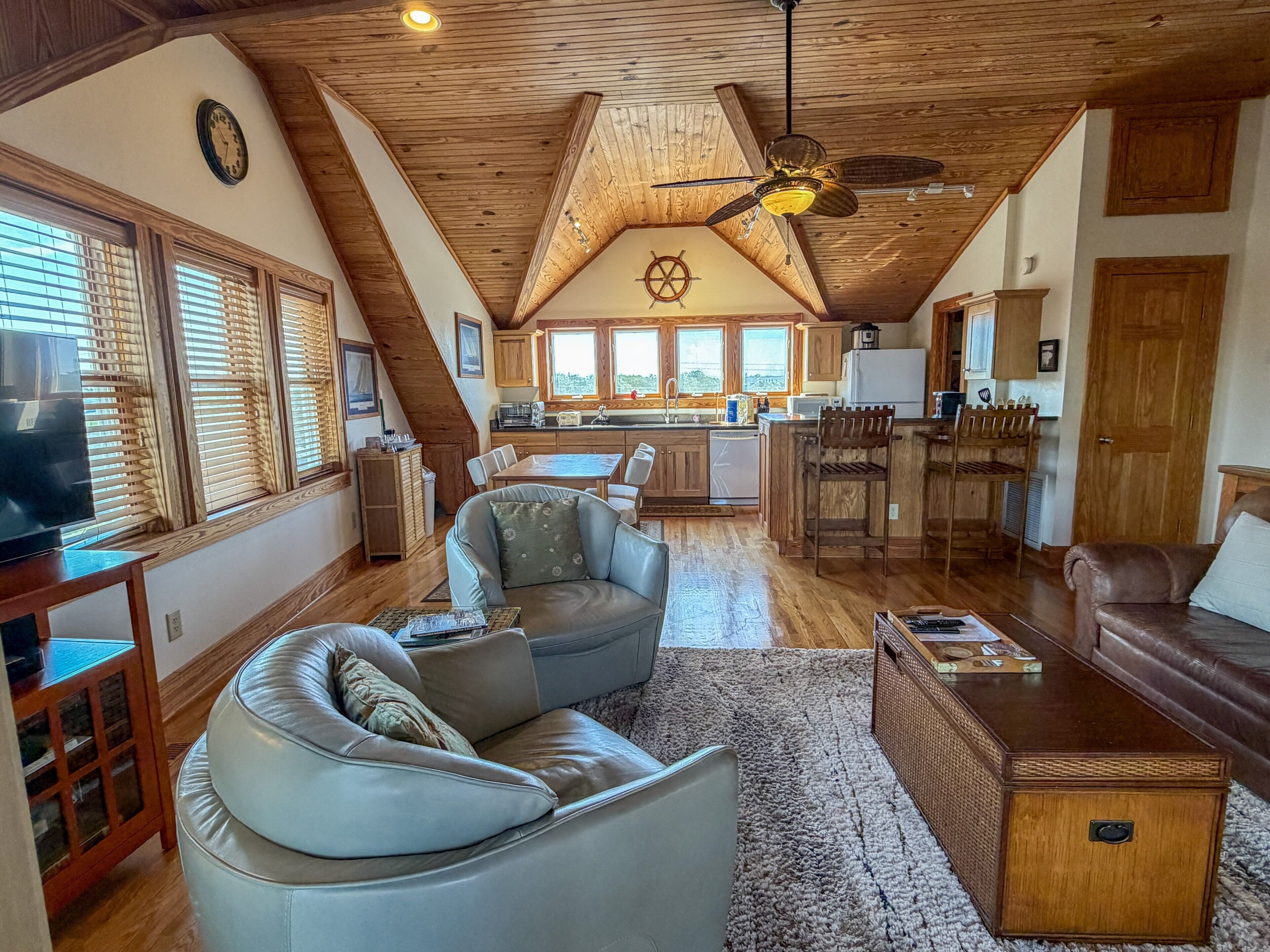 A spacious living area with a high ceiling, wooden beams, a ceiling fan, and a view of the kitchen and dining area.