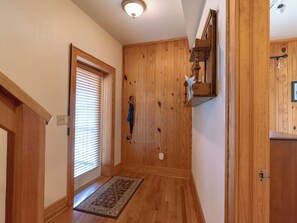 A wooden hallway with a door, a window with blinds, a wall-mounted shelf, and a rug on the floor.