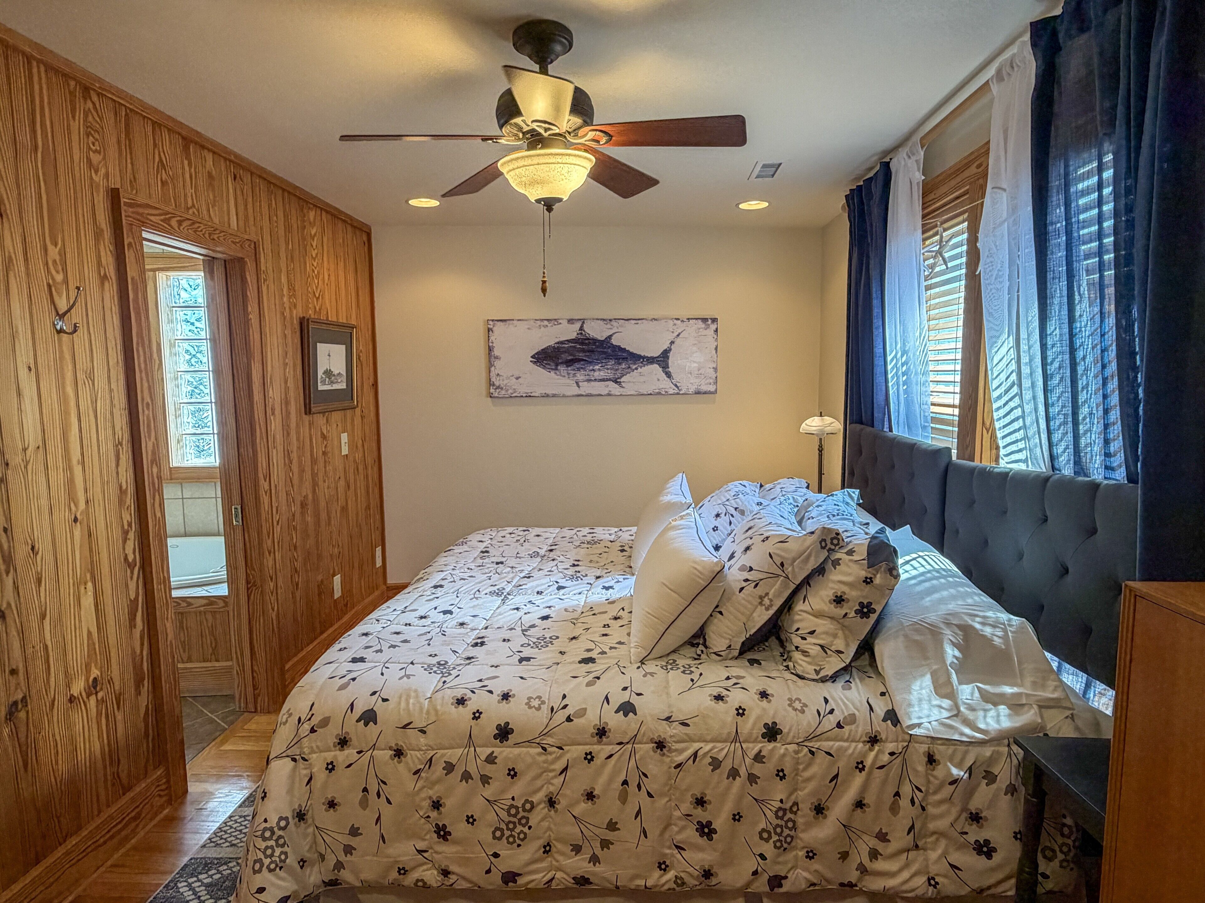 Un dormitorio con cama, ventilador de techo, ventana con cortinas e imagen colgada en la pared.