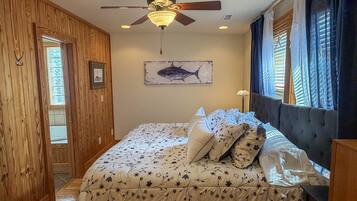 Un dormitorio con cama, ventilador de techo, ventana con cortinas e imagen colgada en la pared.