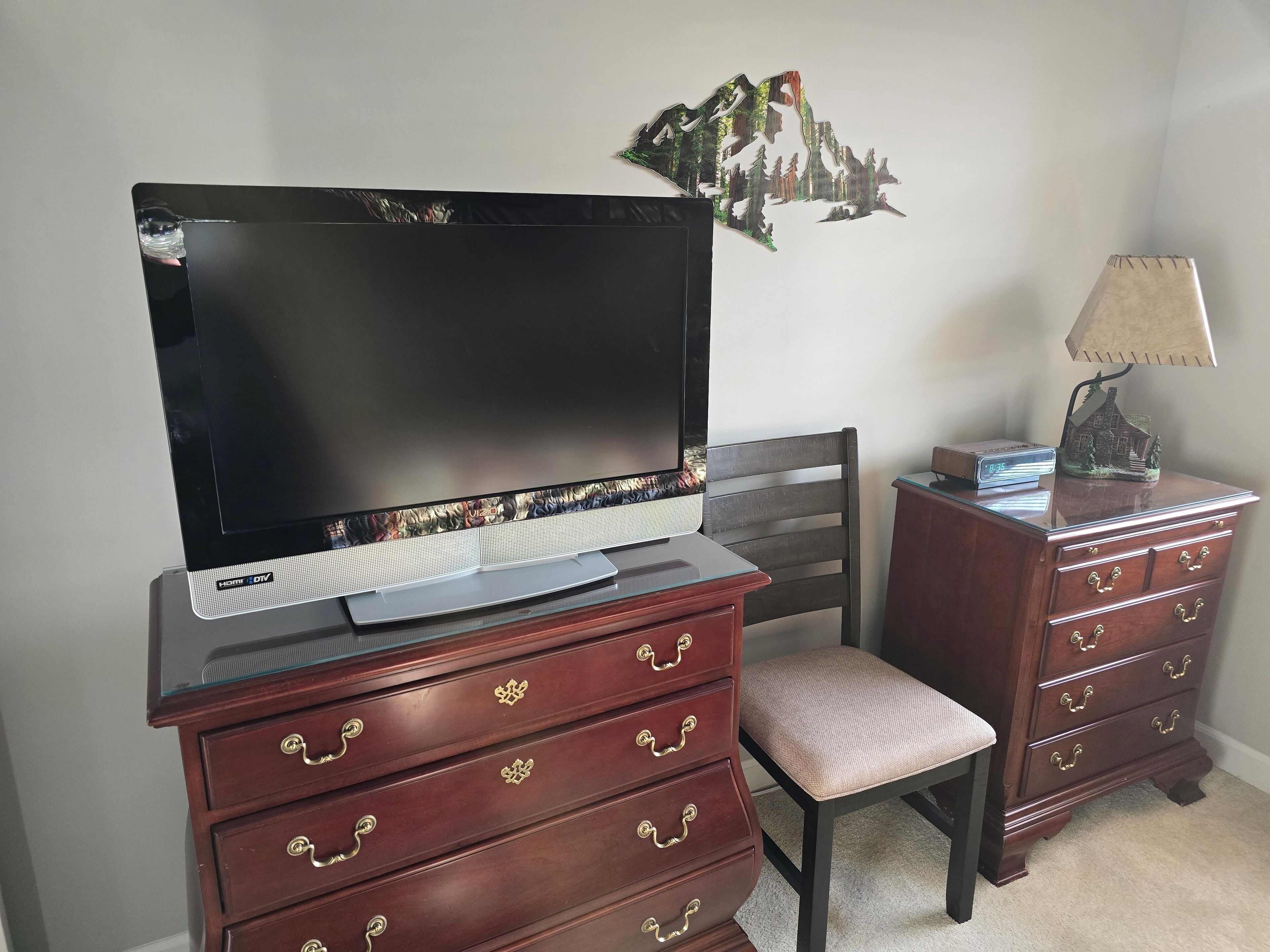 A bedroom with a TV on a stand, a wooden dresser with drawers, a chair, and a lamp on a side table.