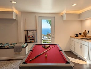 Un tavolo da biliardo con le bocce, una porta che dà su un balcone con vista, e una zona cucina con pensili e lavello.