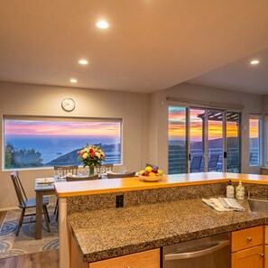 A kitchen with a granite countertop, a dining table with chairs, a vase of flowers, and a view of the sunset through large windows. - Panoramic views 5BR Hot tub golf game room (Malibu)