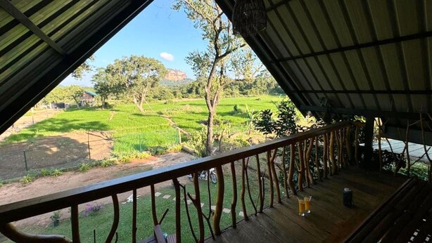 Un espacio al aire libre techado con una terraza de madera, barandilla metálica y vista a un paisaje verde con árboles y campos.