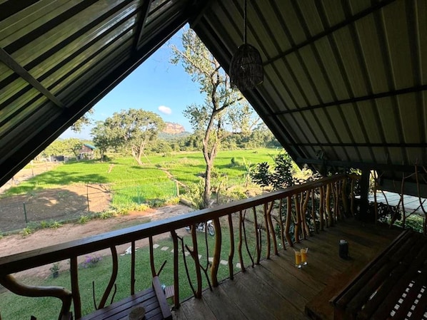 Un espacio al aire libre techado con una terraza de madera, barandilla metálica y vista a un paisaje verde con árboles y campos.