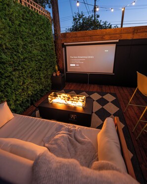 A cozy outdoor seating area with a fire feature, a projector screen displaying 'The Hulu Streaming Library', and a comfortable couch. - Charming bungalow in trendy West Adams with private tranquil outdoor space (Los Angeles)