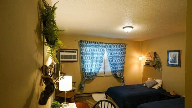 Habitació amb dos llits, un escriptori amb cadira, una làmpada i una finestra amb cortines estampades.