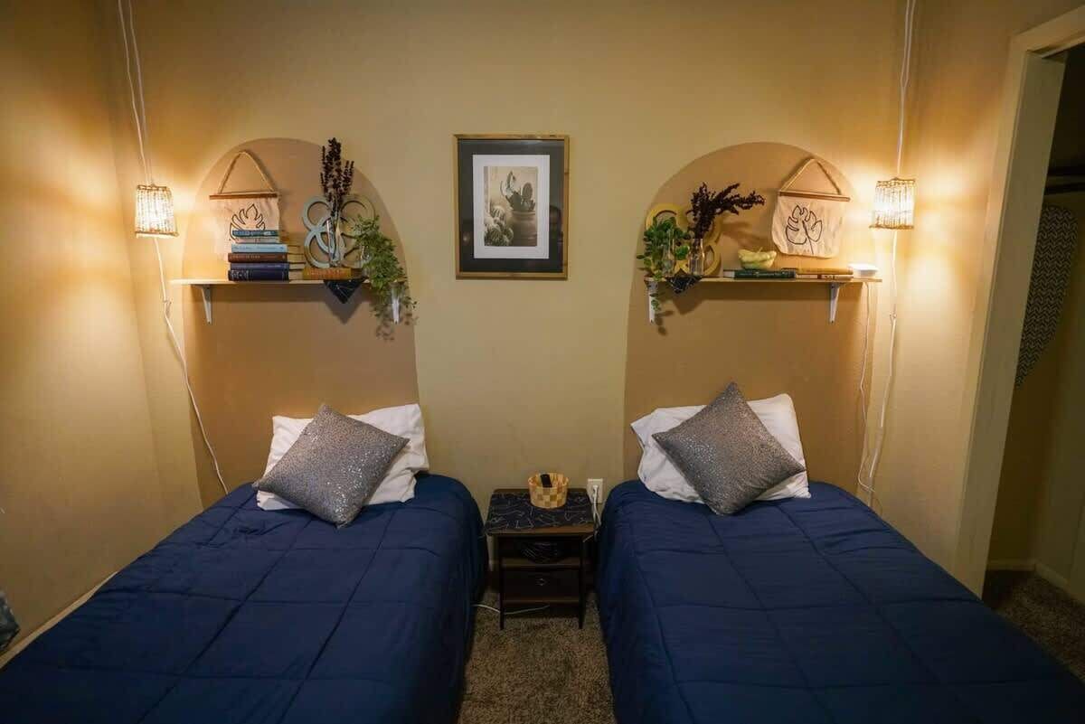 A room with two beds, each with a pillow and a small nightstand. There are wall-mounted shelves with decorative items and a framed picture above the beds.