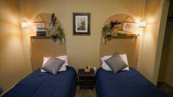 A room with two beds, each with a pillow and a small nightstand. There are wall-mounted shelves with decorative items and a framed picture above the beds.