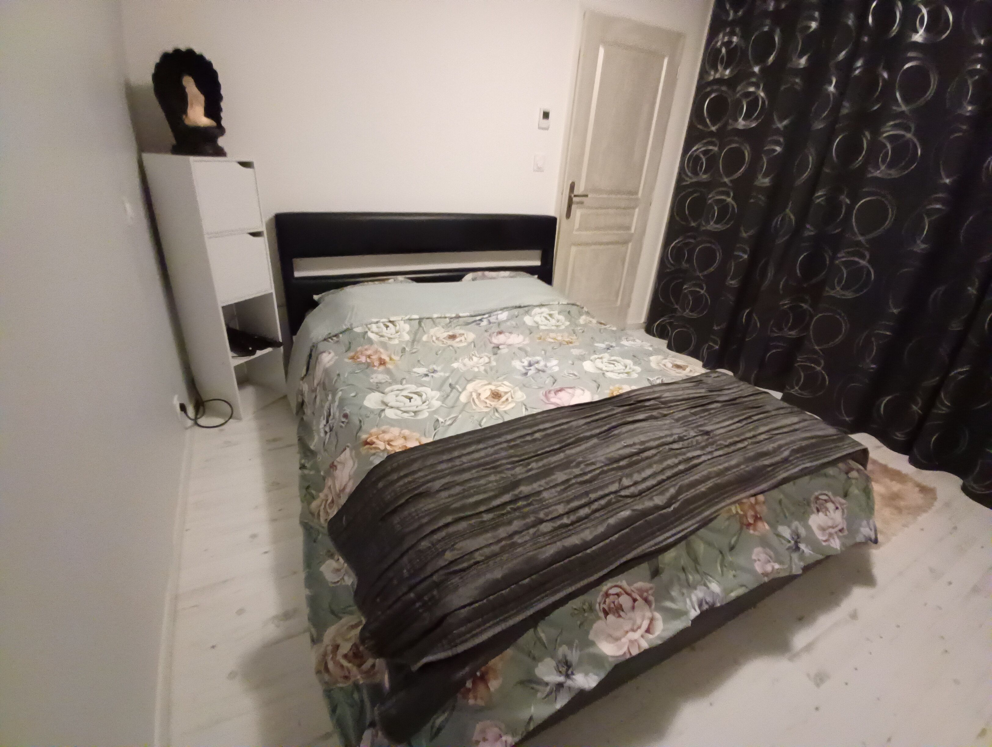 A bed with a floral bedspread and a dark blanket, a bedside table with a lamp, and a door with a circular pattern curtain.