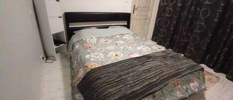 A bed with a floral bedspread and a dark blanket, a bedside table with a lamp, and a door with a circular pattern curtain.