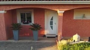 A house with a red facade, a tiled roof, a white door, and a small garden with plants. - House Seasonal rental with swimming pool (Millau)