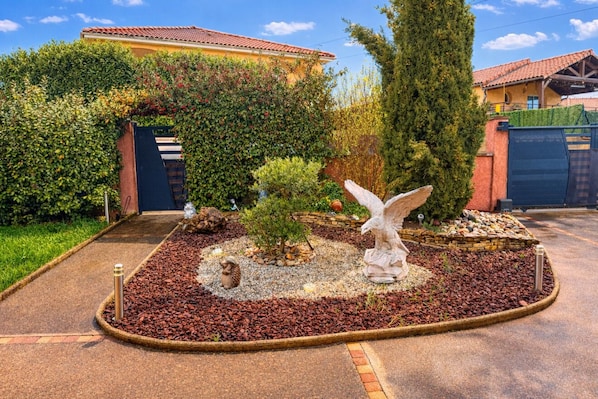 A landscaped garden with a statue, a small tree, and a stone pathway leading to a building with a red-tiled roof. - House Seasonal rental with swimming pool (Millau)