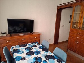 Ein Essbereich mit einem Tisch, der mit einer Tischdecke mit geometrischem Muster bedeckt ist, einem Fernseher auf einer Holzkommode und einer Holzkommode mit Glastüren.