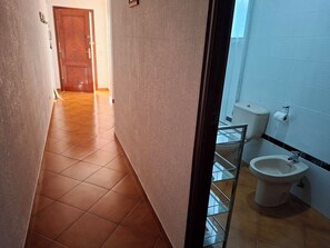 Ein Korridor mit einer Holztür am Ende, der zu einem Badezimmer mit Toilette, Bidet und Fliesenboden führt.