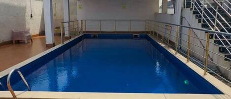 A rectangular swimming pool with a wooden deck surrounding it, a metal railing, and a staircase leading to an upper level.