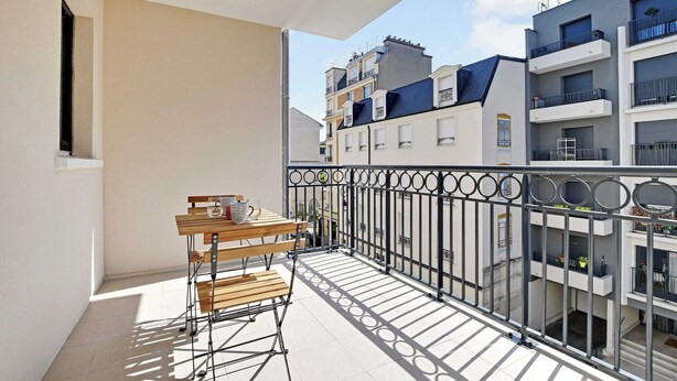 Un balcone con tavolo e sedie in legno, ringhiera metallica e vista sui palazzi limitrofi.